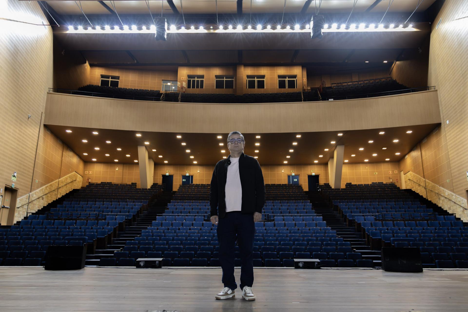 Foto colorida horizontal de Felipe, centralido à frente do palco e da plateia do Centro de Convenções. A foto mostra um homem bem ao centro e detalhes do lugar, como o palco, a plateia e o mezanino