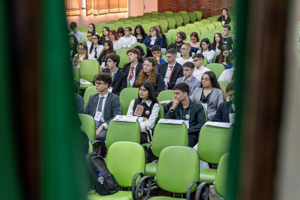 Foto colorida horizontal de um grupo de jovens com roupas formais está sentado em cadeiras verdes, atentos a uma atividade. À esquerda e à direita, partes da imagem estão parcialmente encobertas por cortinas verdes. Ao fundo, mais fileiras de cadeiras vazias e uma parede de tijolos aparentes.