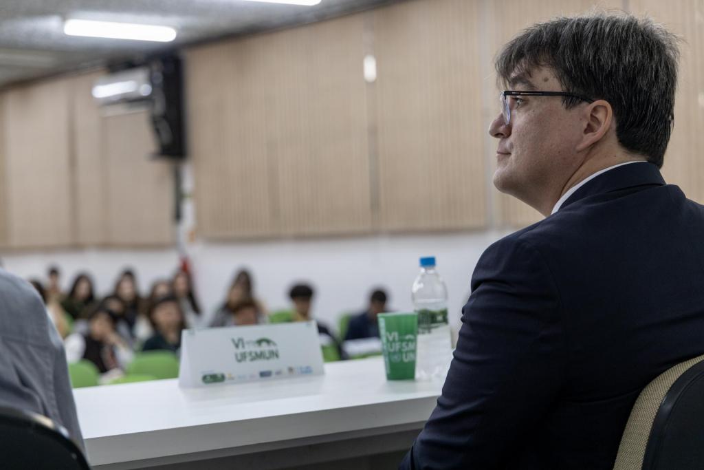 Foto colorida horizontal Homem de terno escuro está à direita, sentado atrás de mesa branca com placa, copo e garrafa de água. Ao fundo, grupo de pessoas com roupas casuais observa a frente da sala.