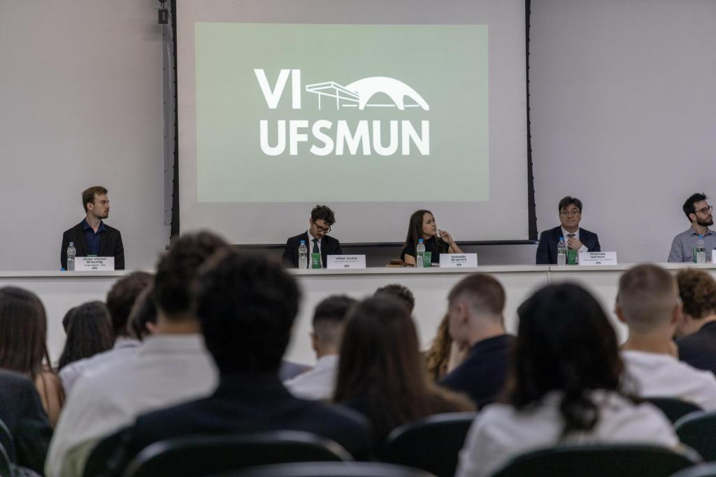 Foto colorida horizontal de cinco pessoas sentadas atrás de uma mesa com garrafas de água, copos e placas de identificação, diante de uma plateia. Ao centro, um telão exibe o logo “VI UFSMUN”. À frente, público com roupas sociais está voltado para o painel.