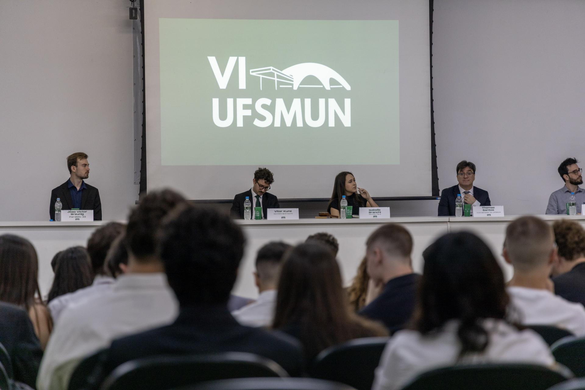 Foto colorida horizontal de cinco pessoas sentadas atrás de uma mesa com garrafas de água, copos e placas de identificação, diante de uma plateia. Ao centro, um telão exibe o logo “VI UFSMUN”. À frente, público com roupas sociais está voltado para o painel.