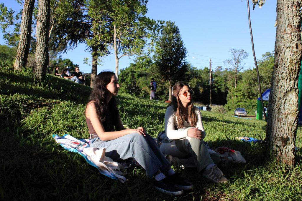 Foto colorida horizontal. Da esquerda para o centro, Maria, uma mulher branca de longos cabelos pretos, está sentada em cima de um pano colocado sobre a grama. Do centro para a direita, Teresa, uma mulher branca de longos cabelos loiros, está sentada do mesmo modo e utiliza óculos de sol. Elas olham para a extremidade direita da foto.