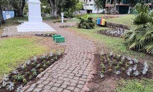 Foto horizontal. Mostra uma praça com canteiros, contendo também árvores, gramado, lixeiras e um monumento pintado de branco