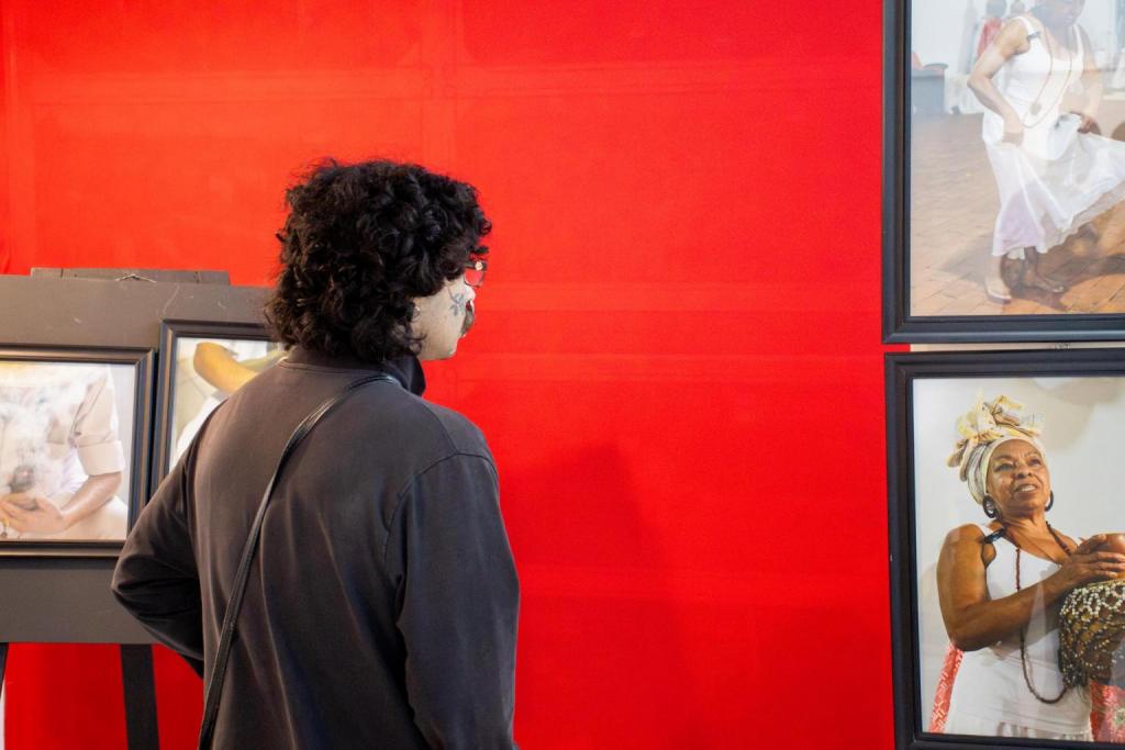 Foto colorida horizontal de homem de costas que observa fotos enquadradas em uma parede vermelha. A imagem mostra parcialmente as fotos de praticantes de cultos afro