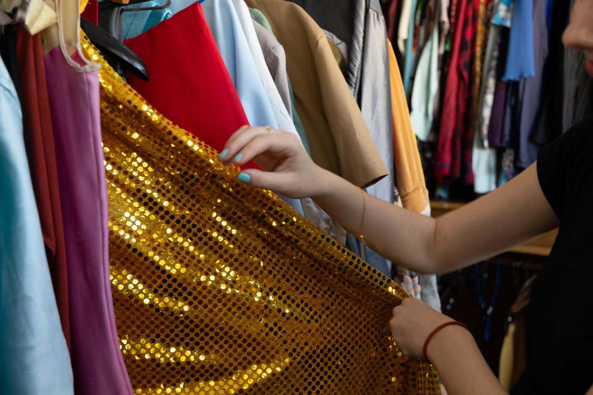 Foto horizontal. Em meio a várias roupas penduradas em cabides, destaca-se um vestido dourado, que é puxado por uma menina branca, cujas unhas estão pintadas de azul claro.
