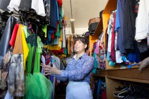 Foto horizontal colorida. A imagem retrata a professora Camila sorrindo e gesticulando em meio a um espaço repleto de roupas penduradas, de diversas cores. No centro da imagem, está uma mulher de cabelos castanhos e camisa lilás estampada, sorrindo e gesticulando com as mãos. Suas unhas estão pintadas de rosa, e a expressão é descontraída.