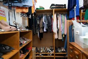 Foto horizontal colorida. O espaço retrata uma parte do laboratório, mostra um armário e prateleiras cheios de roupas e sapatos organizados. Em primeiro plano, no centro da imagem, há um grande armário de madeira com portas abertas, revelando diversas roupas penduradas em cabides. Abaixo delas, sapatos de diferentes estilos estão dispostos lado a lado em prateleiras. Sobre o móvel, aparecem algumas malas empilhadas. À esquerda, há parte de outra estante com mais roupas coloridas. A luz natural penetra suavemente pela lateral, destacando os diferentes tons de tecido. Ao fundo, é possível ver mais roupas penduradas e parte da parede branca do ambiente, completando a sensação de um acervo de figurino bem organizado.