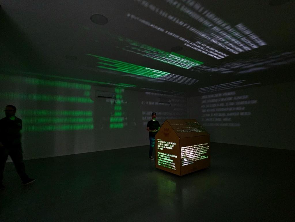 Foto colorida horizontal de sala escura com projeções em textso em verdes no teto e nas paredes. No canto direito da sala escura, um homem em uma mesa controla as projeções