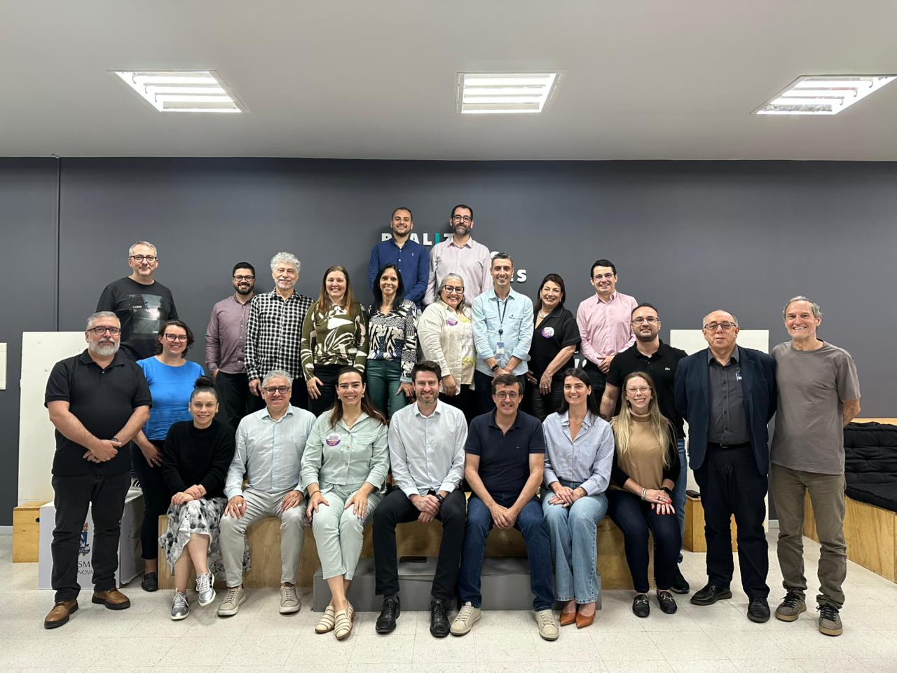 foto colorida horizontal de um grupo de pessoas, alguns sentados e outros em pé, em pose oficial, com uma parede chumbo ao fundo