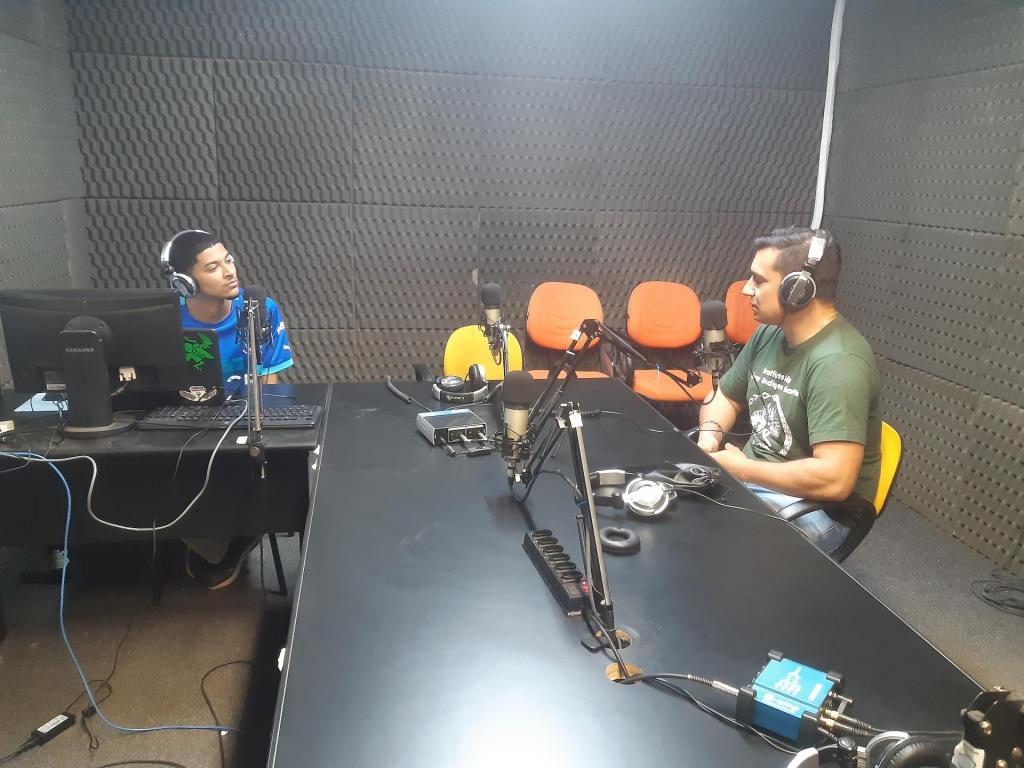 Foto colorida horizontal de duas pessoas em um estúdio de rádio. Do lado esquerdo, o estudante Felipe, que usa um azul e um fone de ouvido. Ele está sendo atrás de um monitor de computador. Do lado direito, sentado de lado, o professor Willian, que vest uma camiseta verde e usa fones de ouvido.