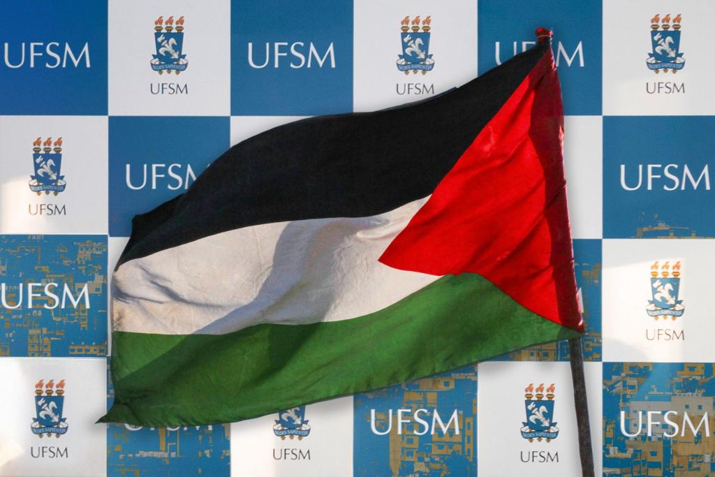 Foto retangular, horizontal e colorida de uma bandeira da Palestina em frente a um mural com a identidade visual da UFSM.