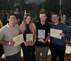 Foto colorida e horizontal de quatro jovens que estão lado a lado, sorrindo e segurando prêmios. Da esquerda para a direita: um homem de óculos veste camisa bege e segura um certificado, uma mulher de cabelo loiro veste vestido preto e segura um troféu com mosaico colorido, outro homem, também segura um troféu semelhante e veste camisa preta e o quarto homem, de óculos e bigode, veste camisa azul e segura um certificado. Ao fundo, várias pessoas estão em pé, em um ambiente iluminado, com árvores e decoração de natureza.