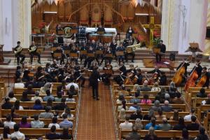 Foto horizontal: A imagem aérea mostra os músicos da orquestra (todos eles vestidos de preto) tocando seus instrumentos em frente ao altar da catedral. O público está sentado nos bancos da igreja.