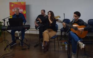Foto horizontal. Tendo ao fundo uma parede branca, quatro músicos estão sentado em cadeiras azuis. Há um tecladista, um baixista, um violonista e uma cantora.