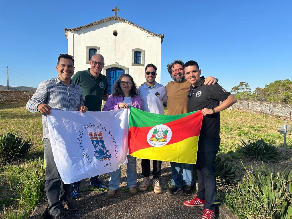 Foto colorida horizontal de seis pessoa, sendo cinco homens e uma mulhes. Todos usam roupas casuais e seguram as bandeiras da UFSM e do Rio Grande do Sul. Atrás deles, uma pequena igreja branca. A imagem foi feita em um dia com tempo firme.