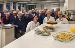Foto horizontal. À direita, está uma mesa branca, sobre a qual estão quatro pratos contendo diferentes alimentos, além de um copo com suco de laranja. À frente da mesa, estão cerca de 20 pessoas, todas elas usando toucas brancas de cozinha na cabeça. Três mulheres vestem uniforme branco e os demais, uniforme azul escuro.