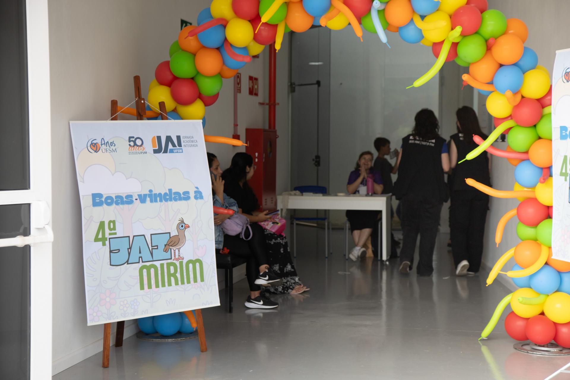 Foto colorida horizontal de entrada de prédio. No alto e dos lado, uma cachopa de balões. Do lado esquerdo, um banner da 4ª JAI Mirim. Ao fundo, mesas de credenciamento