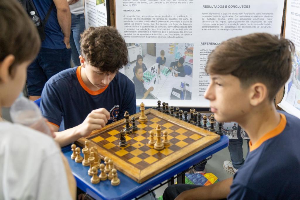 Foto colorida horizontal de dois meninos jogando xadrez. O menino que está do lado esquerdo está com a mão levantada em movimento direcionado ao tabuleiro. O menino da direita está com o olhar voltado para lado contrário ao tabuleiro. No tabuleiro, poucas peças. Dos lados, peças brancas e pretas.