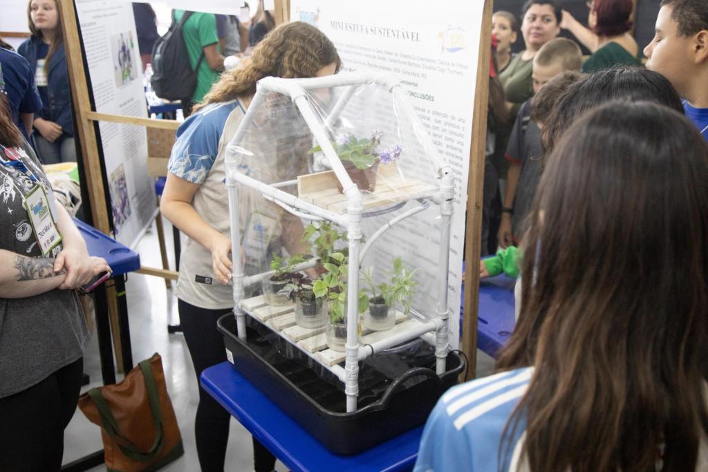 Foto colorida horizontal de pequena estufa criada por estudantes. A estrutura pequena branca tem algumas plantas. Atrás da estufa, uma menina organiza as plantas. No entorno, outras crianças