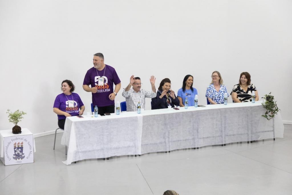 Foto colorida horizontal de sete pessoas atrás da mesa de um evento. Do grupo, seis estão sentados e um está em pé . Ainda, o terceiro sentado, o professor Flavi, está com as mãos levantandas. Todos estão sorrindo.