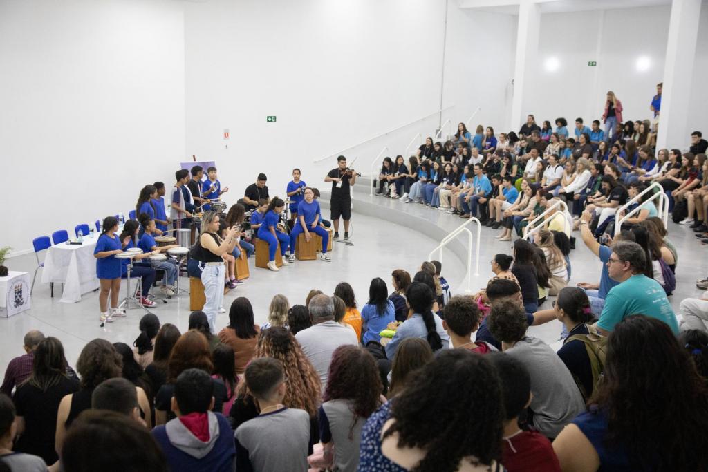 Foto colorida horizontal de grupo de percursão se apresentando ao centro do Museu do Conhecimento. Além de instrumentos de percussão, o grupo tem um violinista. A platéia está posicionada na arquibanca circular