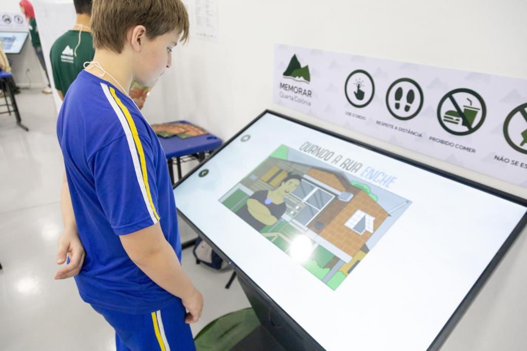 Foto colorida horizontal de menino posicionado ao lado esquerdo. Ele acessa um totem digital que está inclinado e mostra a imagem da HQ Quando a rua enche