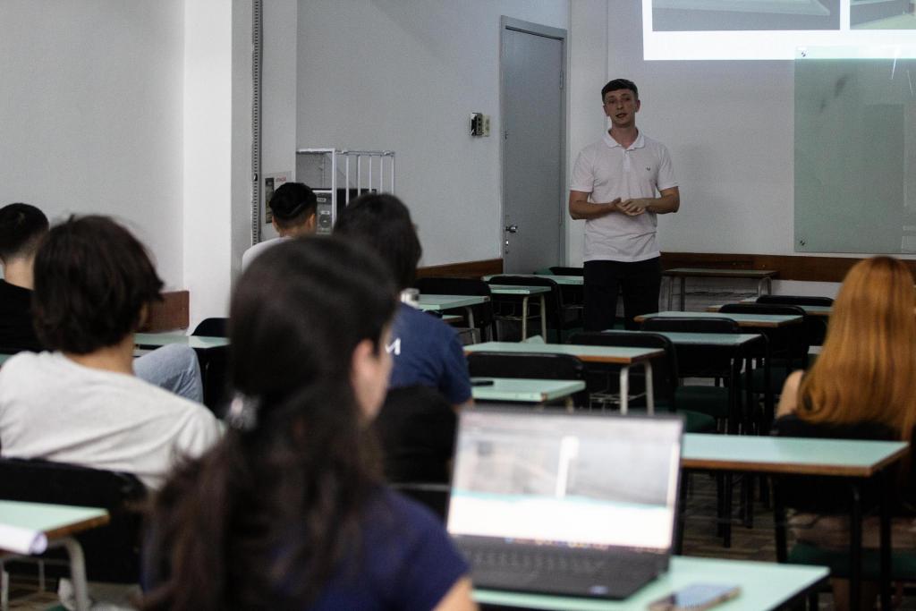 Foto colorida horizontal de apresentação de trabalho. Ao fundo, um universitário em pé apresenta trabalho. À frente dele, grupo de estudante sentados em classes escolares. Alguns deles estão com notebook ligado.