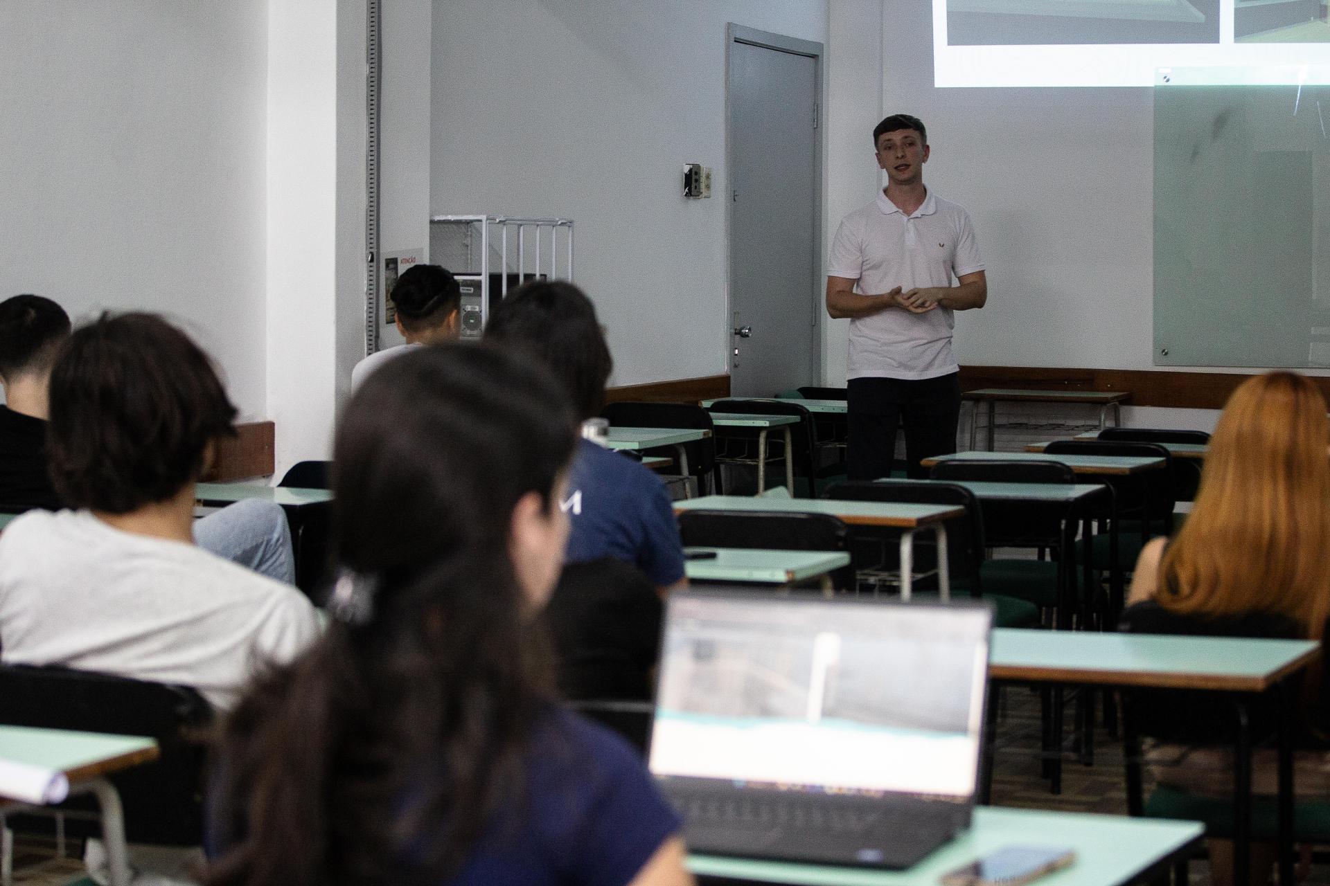 Foto colorida horizontal de apresentação de trabalho. Ao fundo, um universitário em pé apresenta trabalho. À frente dele, grupo de estudante sentados em classes escolares. Alguns deles estão com notebook ligado.