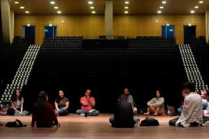 Foto horizontal colorida. Mostra um grupo de pessoas sentadas em círculo sobre o palco de um auditório vazio. Em primeiro plano, pessoas estão descalças ou com calçados leves, formando uma roda de conversa no centro do palco de madeira. Ao fundo, as fileiras de assentos escuros do auditório permanecem vazias e iluminadas pelas luzes dos corredores laterais. A iluminação superior destaca o grupo central, sugerindo uma atividade de reflexão, ensaio ou oficina artística. À esquerda, uma mulher de blusa branca e calça verde; ao centro, várias pessoas em posição relaxada, incluindo um homem de camiseta vermelha e outro de blusa colorida; à direita, mais participantes, alguns de costas e outros atentos à conversa.