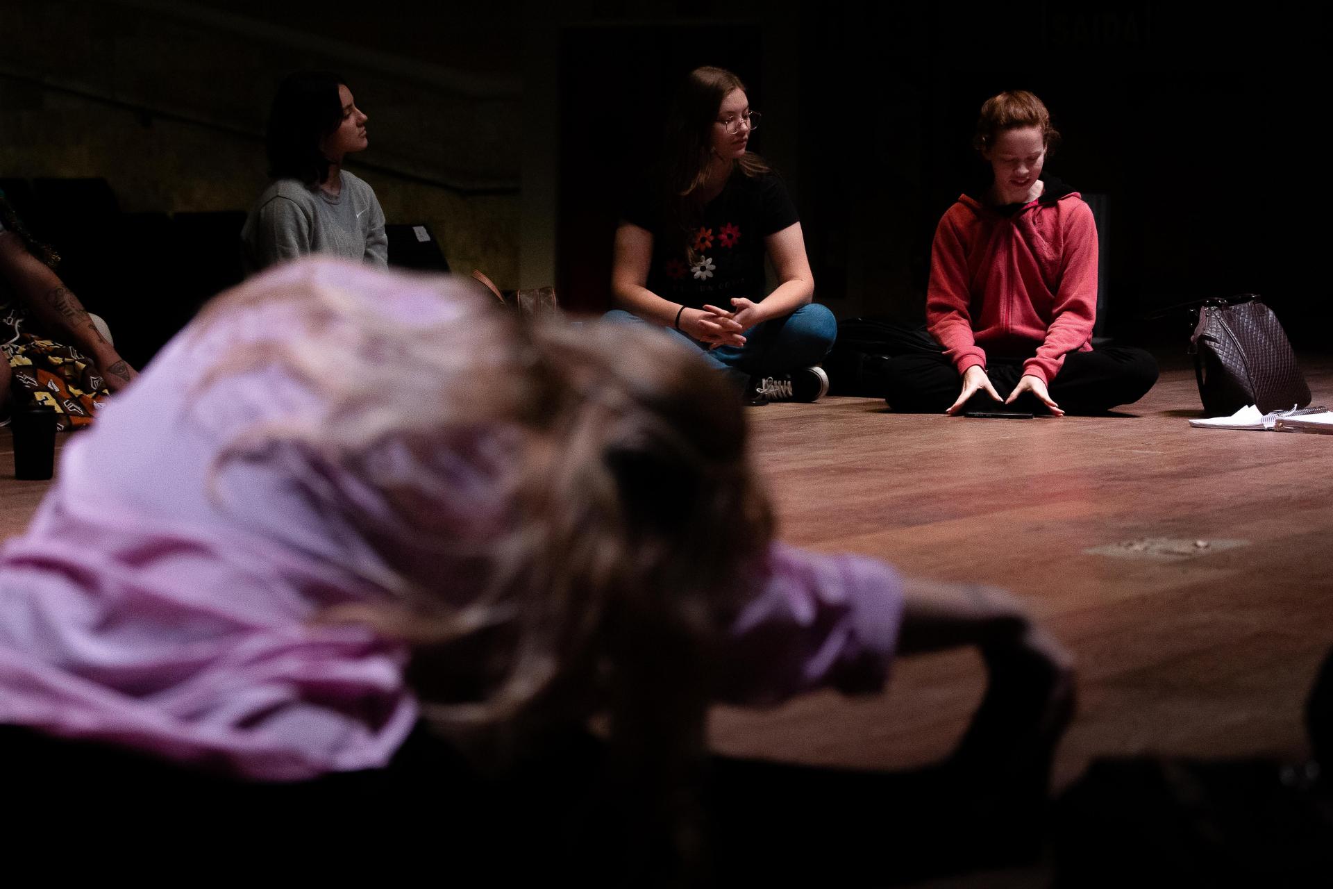 Foto horizontal colorida. A imagem mostra um grupo de mulheres sentadas no palco. Uma delas, de costas, faz alongamento. A iluminação é suave e concentrada nelas, destacando expressões de atenção e concentração.
