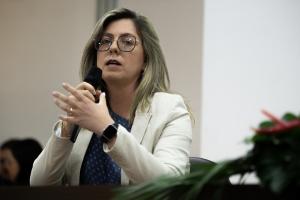 Uma mulher está sentada, segurando um microfone e falando. Ela é branca, usa óculos, tem cabelo loiro médio, usa uma blusa azul, casaco branco, uma pulseira e um anel.