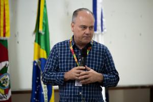 Um homem está em pé falando ao microfone, a foto é tirada mostrando apenas a parte superior do seu corpo. Ele é branco, usa um crachá com cordão amarelo, verde e vermelho, e uma camisa xadrez azul e branca; Atrás dele é possível ver as bandeiras do Brasil e do Rio Grande do Sul.