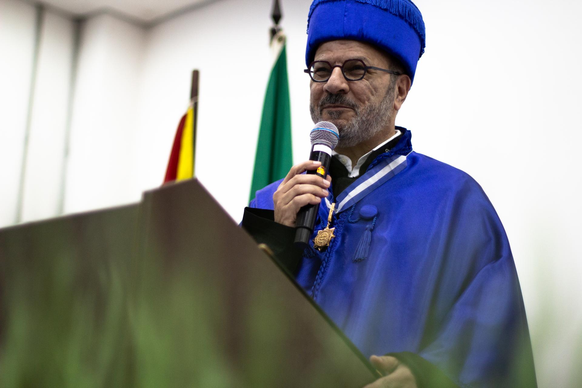Frente a um fundo branco, onde se entreveem bandeiras do Brasil e do Rio Grande do Sul, um homem em pé segura um microfone. Ele é branco, tem barba grisalha e usa óculos, e veste uma beca formal universitária de cor azul, a qual inclui um capelo sobre a cabeça.