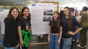 Quatro meninas posam para uma foto, duas de cada lado, em frente a um banner, preso a um suporte de madeira. Ao fundo, há outras pessoas, e também outros banners em suportes de madeira.