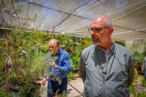Foto horizontal. Na foto, aparecem dois homens. Um veste uma camisa azul e outro, um casaco cinza. Eles estão rodeados por plantas de espécies variadas.