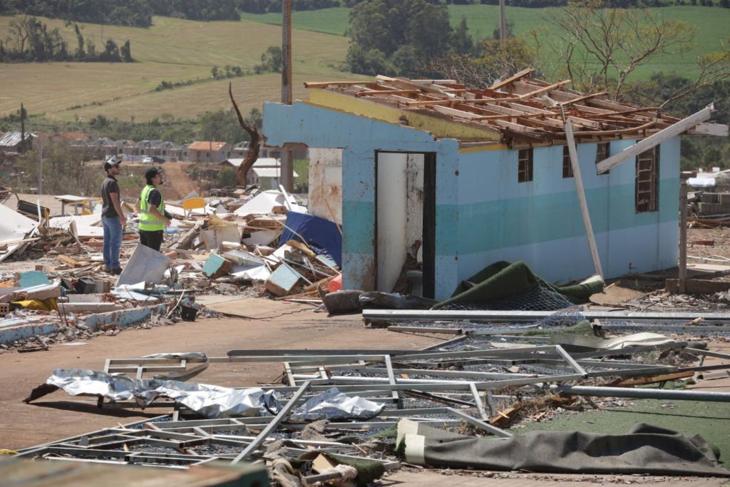 Foto horizontal e colorida de uma área totalmente devastada, com escombros espalhados por todo o chão. No centro, há uma construção parcialmente de pé, com paredes azul-claro e sem telhado. Dois homens, um usando colete refletivo de capacete, e outro de boné, observam os danos enquanto caminham pelo local. Ao fundo, é possível ver um bairro rural e campos agrícolas, reforçando a dimensão da destruição.