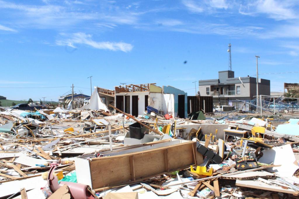 Foto horizontal e colorida de um cenário de destruição extrema: restos de casas, móveis, estruturas metálicas e telhados estão espalhados pelo chão. Uma fileira de portas indica o que antes parecia ser um bloco de residências ou salas. Ao fundo, prédios ainda de pé contrastam com a área totalmente demolida. A paisagem evidencia a força do evento climático que atingiu a região.