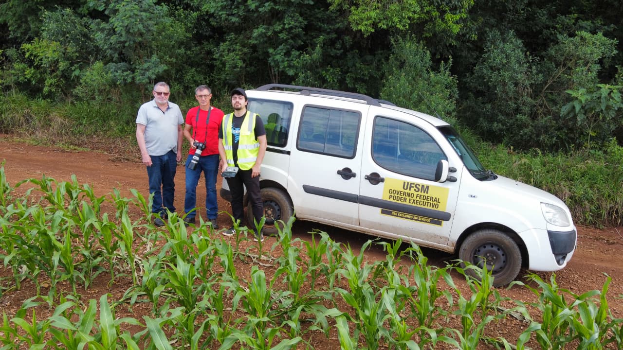 Na foto, três homens posam ao lado de um veículo branco da UFSM, estacionado em uma estrada de terra cercada por vegetação. Um deles veste colete refletivo e segura um equipamento, sugerindo atividade de campo ou operação de drone. Os outros dois carregam câmeras, indicando registro técnico ou jornalístico. A cena retrata parte da equipe enviada para avaliação dos impactos do desastre.
