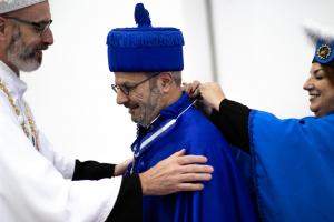 Três pessoas participam de uma cerimônia acadêmica formal. No centro, está um homem de meia-idade, com barba grisalha e óculos, vestindo uma capa e um chapéu cerimonial azul intenso, com franjas na borda e um adorno no topo. Ele sorri discretamente enquanto recebe uma honraria. À esquerda, outro homem, também de barba grisalha e óculos, veste uma roupa cerimonial branca com detalhes dourados. Ele segura os ombros do homem ao centro de maneira acolhedora, como parte do ritual. À direita, uma mulher com cabelo castanho veste uma capa cerimonial azul semelhante. Ela sorri enquanto prende uma insígnia ou colar no traje do homem ao centro. O clima da cena é solene e caloroso, típico de cerimônias universitárias.