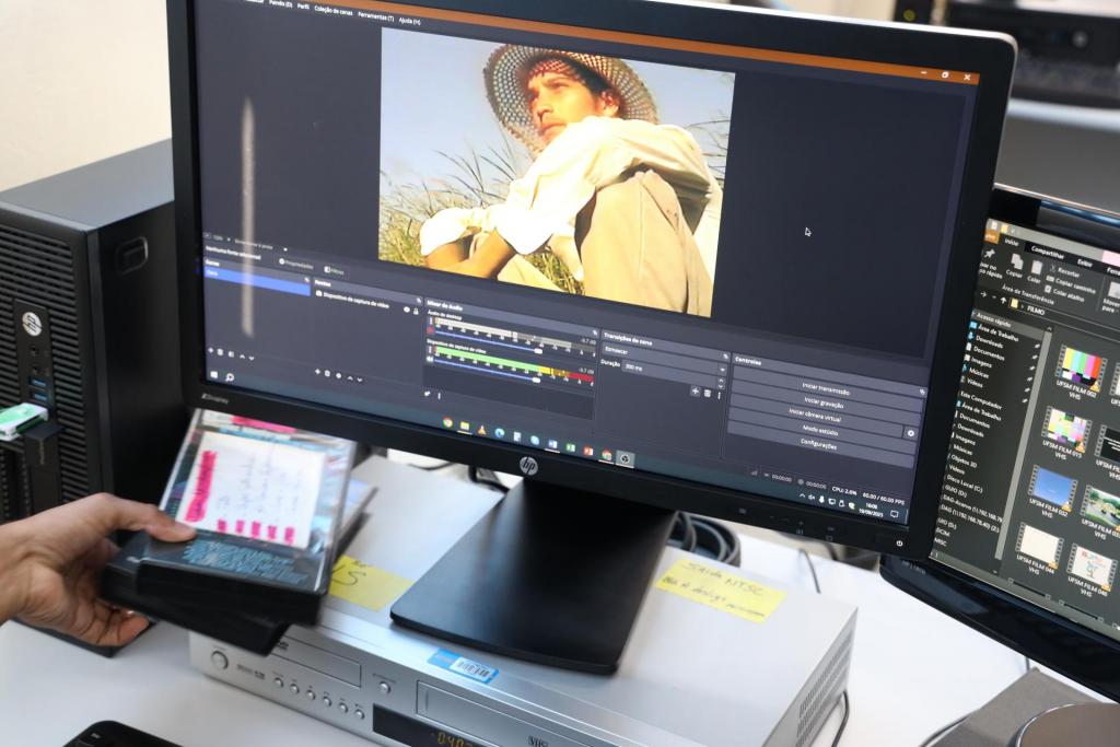 Foto colorido horizontal de monitor de vídeo com uma imagem de um programa de edição com foto enquadrada de um homem com chapéu de palha. A tela também mostraAo fundo, outro monitor ligado. No canto esquerdo, uma mão segura um caixa de fita VHS