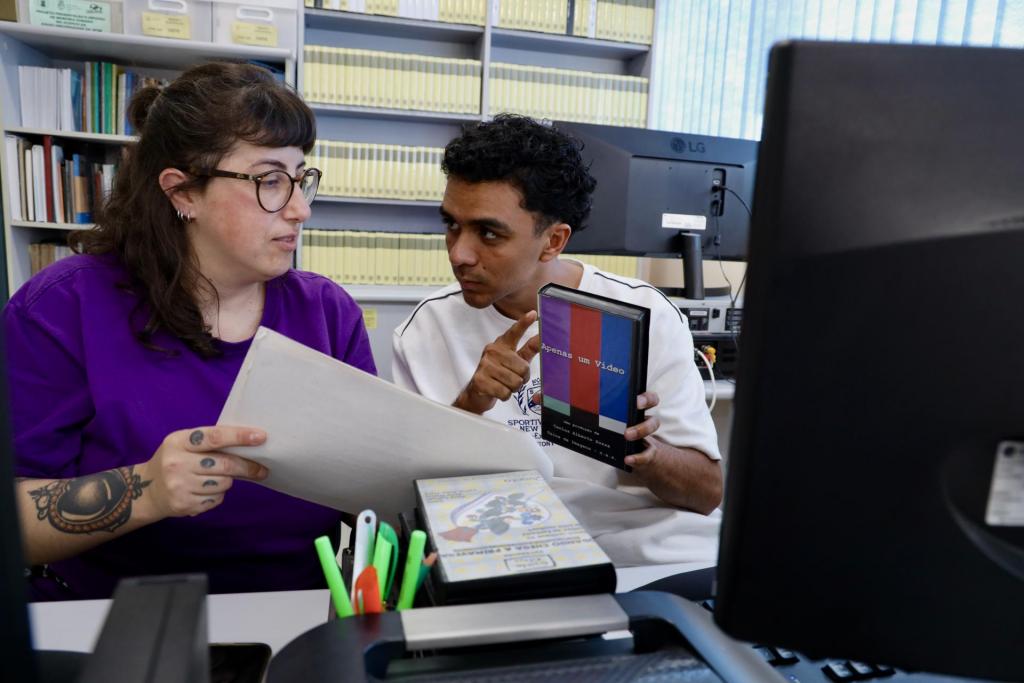 Foto colorida horizontal de mulher e homem atrá de um computador e à frente de uma estante cheia de fitas de vídeos. A mulher usa uma blusa roxa e óculos. Ela segura uma folha. O homem, com uma camiseta branca, segura uma fita de vídeo