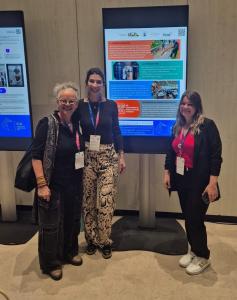 Foto vertical. Na imagem, aparecem três mulheres em pé, tendo ao fundo um pôster apresentado digitalmente em uma tela.