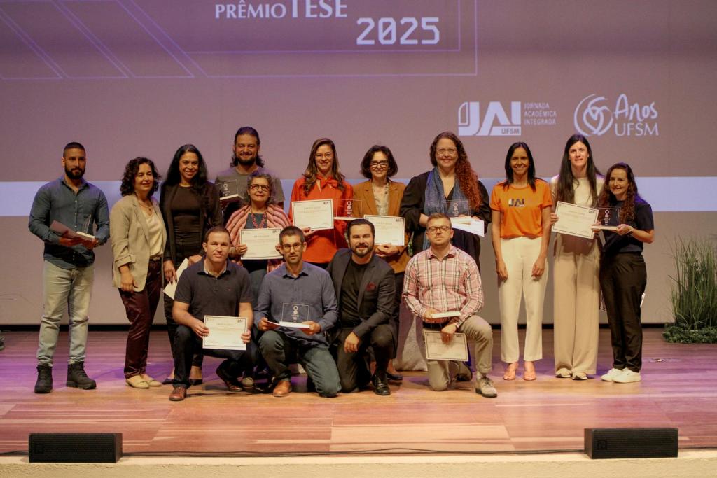 Foto colorida horizontal de grupo de pessoas no palco do Centro de Convenções. O grupo é formado por 14 pessoas, entre homens e mulheres de difentes faixas etárias e fenótipos. Quatro homens estão à frente, agachados. Atrás, os demais em pé. Todos seguram certificados em papel. Atrás do grupo, uma projeção com a imagem do Prêmio Tese
