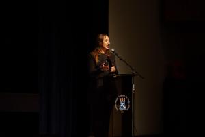 Foto horizontal. Uma mulher branca de cabelos castanhos longos, vestida de preto, fala ao microfone. Ela aparece sobre um fundo escuro e, em frente dela, há um púlpito com o brasão da UFSM