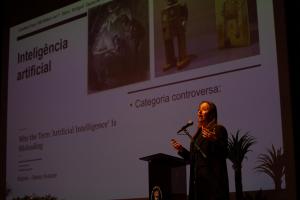 Foto horizontal. No canto direito inferior da imagem, aparece uma mulher branca de cabelos castanhos longos, vestida de preto, que está falando ao microfone. Ao fundo, aparece um slide sobre inteligência artificial, tema de sua palestra.