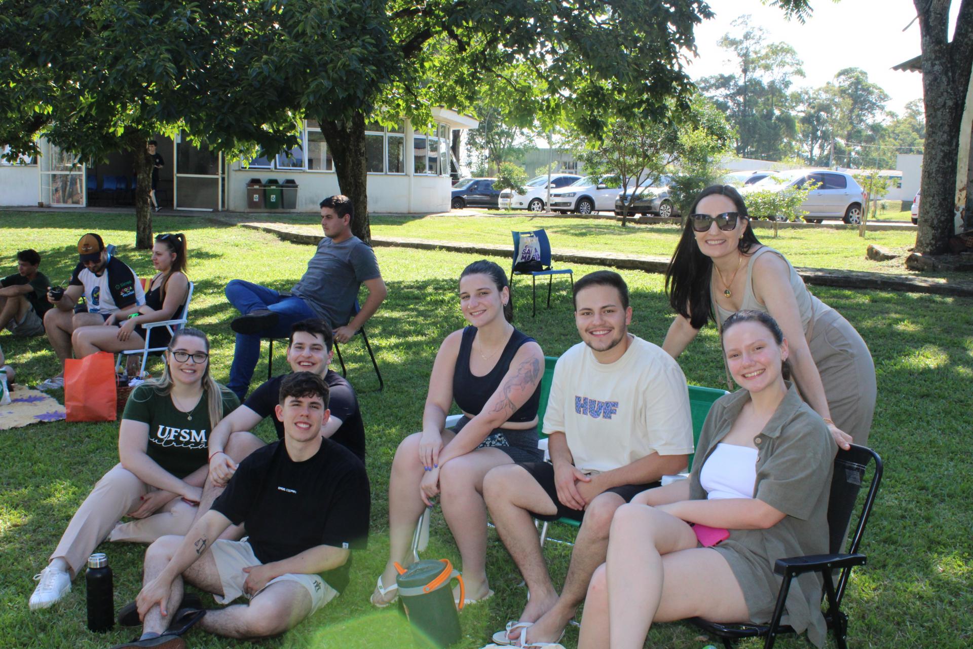 Foto colorida horizontal de grupo de jovens, mulheres e homens, sentados em em cadeiras de abrir e no gramado