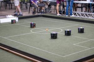 Foto horizontal. A imagem mostra uma reprodução de um campo de futebol, semelhante a uma mesa de futebol de botão, na qual aparecem cinco pequenos robôs em formato cilíndrico. Uma pequena bola está posicionada no centro da cancha.