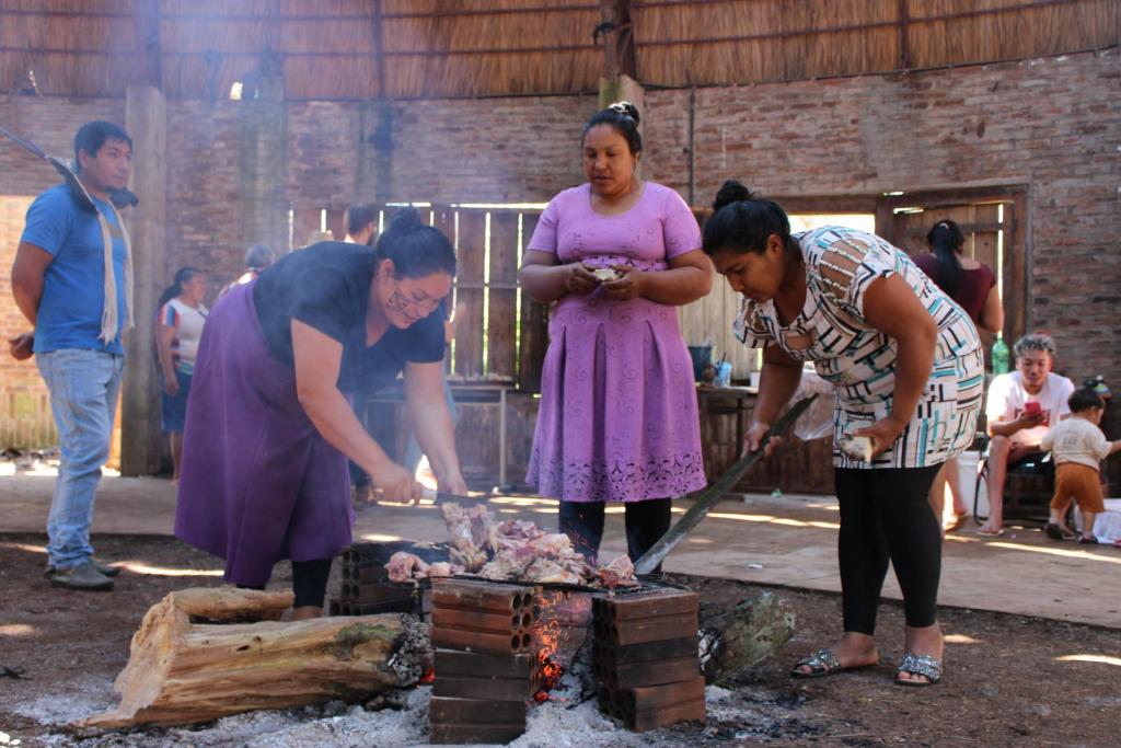 Foto colorida horizontal. Ao centro, indígenas Kaingang preparam a carne numa fogueira cercada por tijolos.