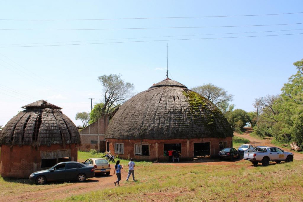 Foto colorida horizontal. Ao centro, o tronco alto do que já foi uma árvore sustenta a estrutura Kaingang central. Feixes de luz passam pelo teto circular, que não está totalmente coberto.