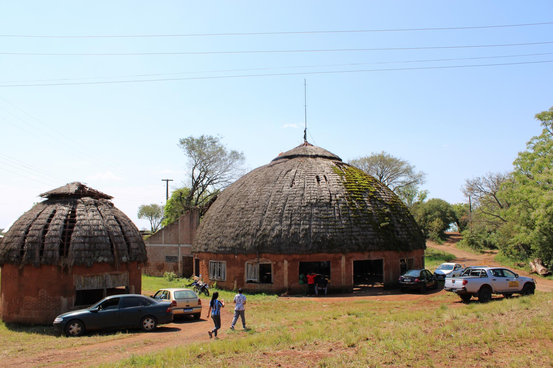 Foto colorida horizontal. Ao centro, o tronco alto do que já foi uma árvore sustenta a estrutura Kaingang central. Feixes de luz passam pelo teto circular, que não está totalmente coberto.